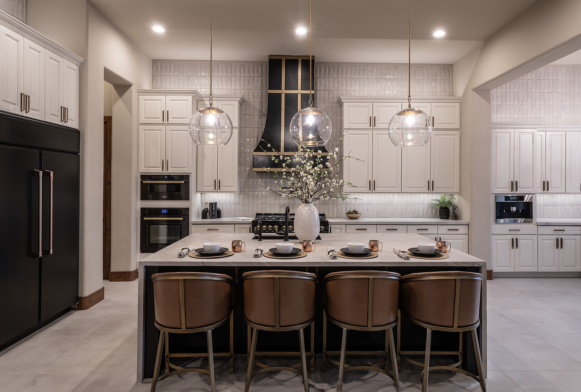 A modern kitchen with white cabinets, a central island with five leather stools, three pendant lights, and stainless steel appliances. A vase with flowers decorates the island.