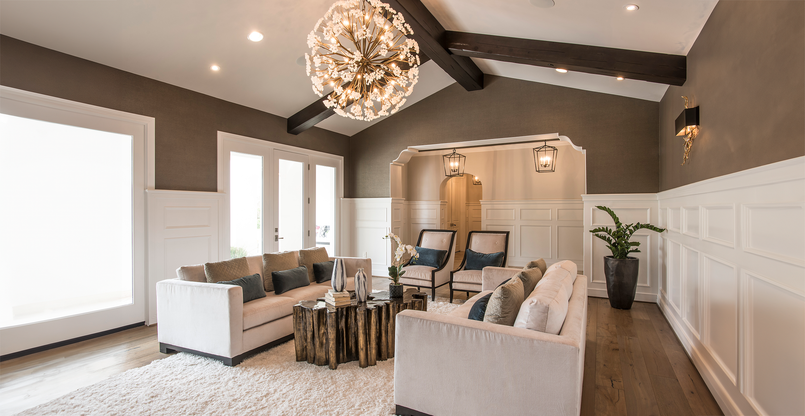 Modern living room with neutral tones, featuring a large chandelier, exposed dark beams, two beige sofas, and wooden coffee table. The room has white wainscoting, recessed lighting, and decorative plants.
