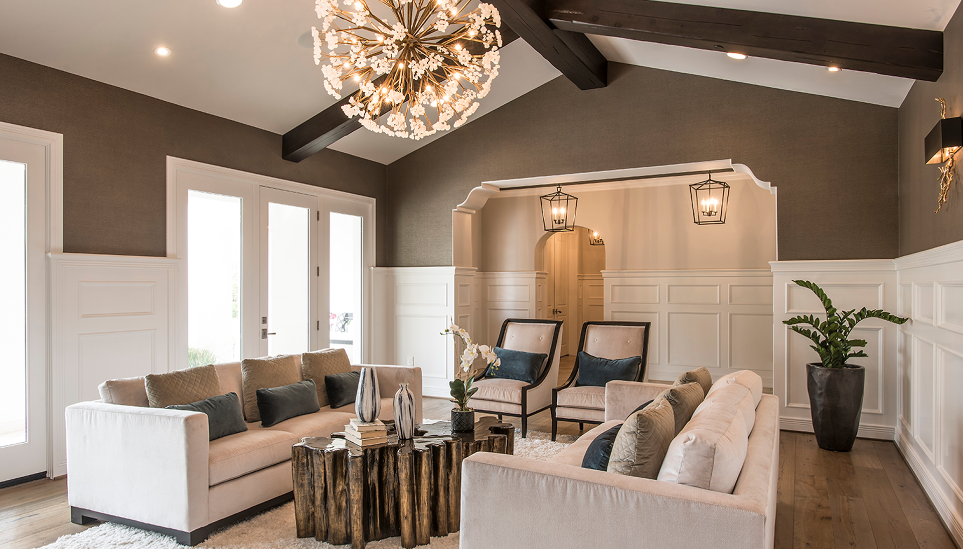 Modern living room with neutral tones, featuring a large chandelier, exposed dark beams, two beige sofas, and wooden coffee table. The room has white wainscoting, recessed lighting, and decorative plants.