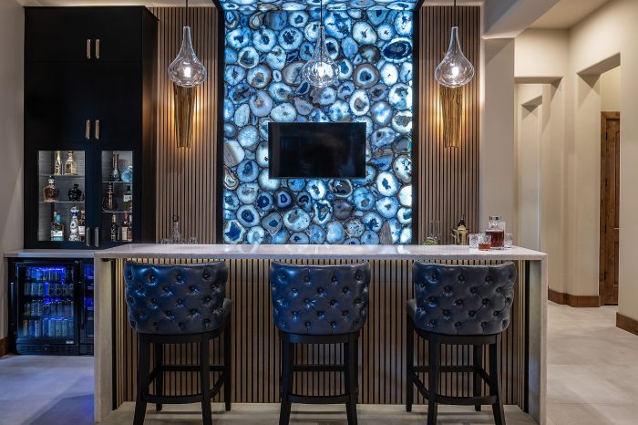 Bar with backlit wall design, mounted T.V., three leather barstools at island and hanging lights.